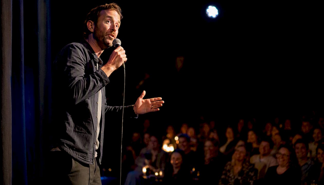 En kvinna står på scen och kör standup inför publiken.
