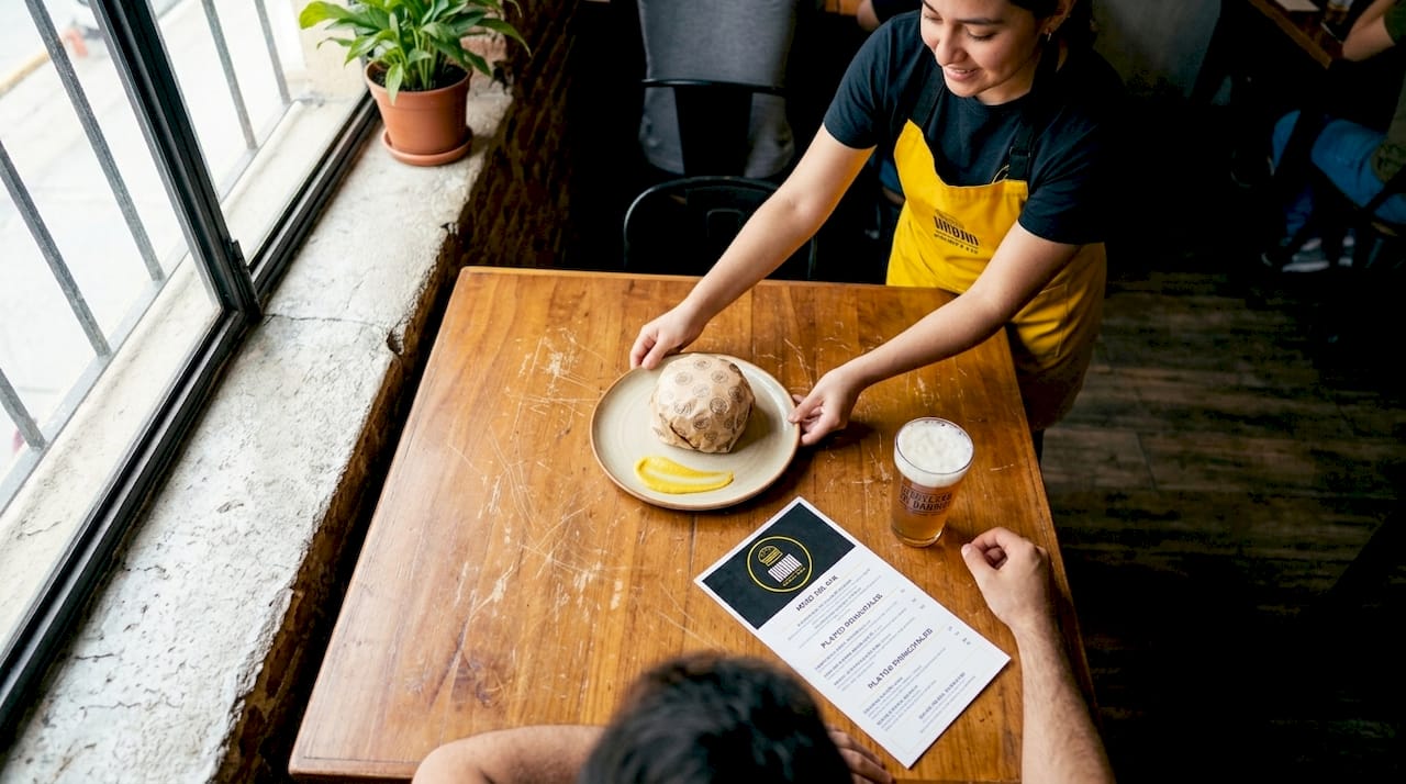 Una camarera lleva a la mesa una hamburguesa recién hecha acompañada de una cerveza artesanal.