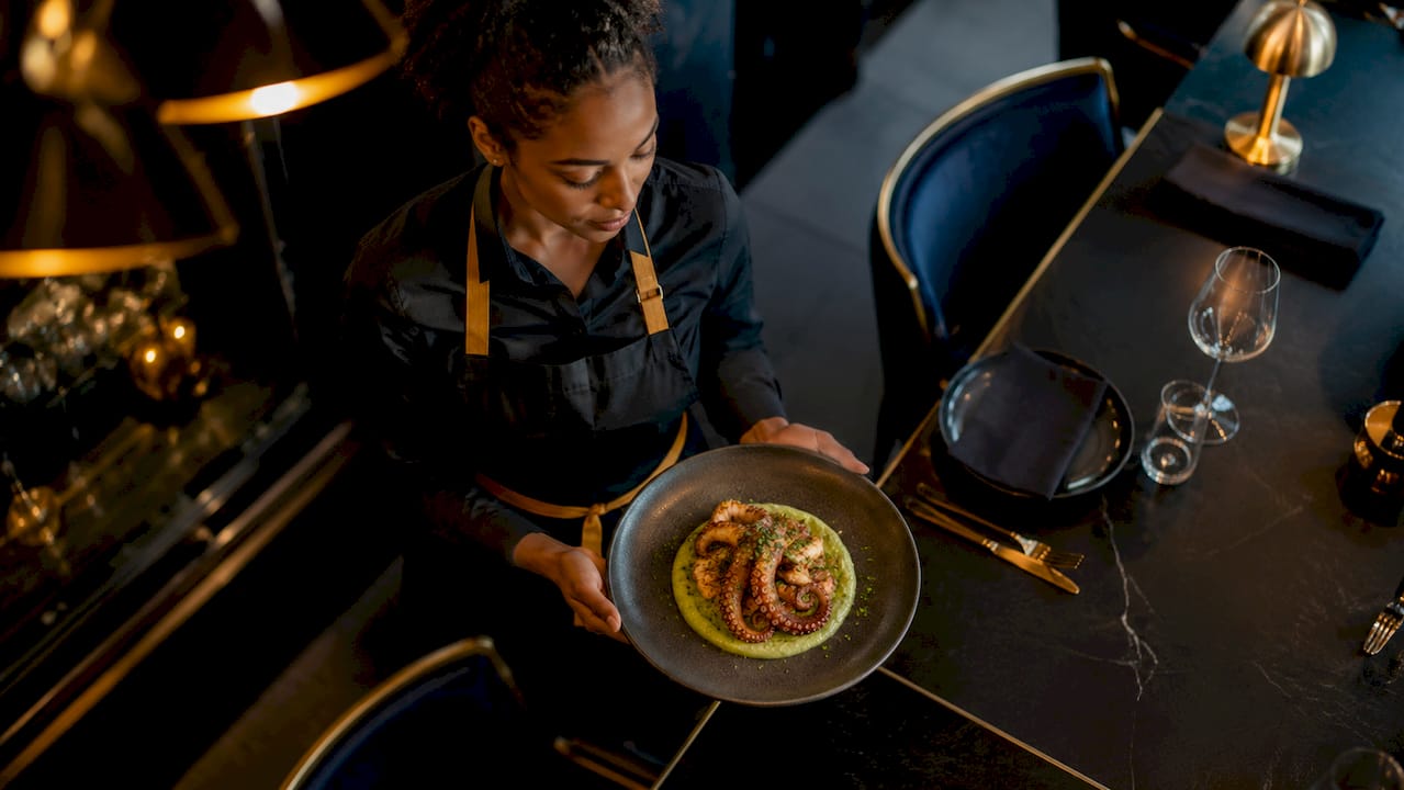 Una camarera presenta un plato de pulpo con una presentación original y llamativa.