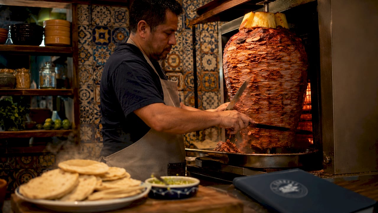 Un taquero cortando carne del trompo para preparar unos tacos al pastor.