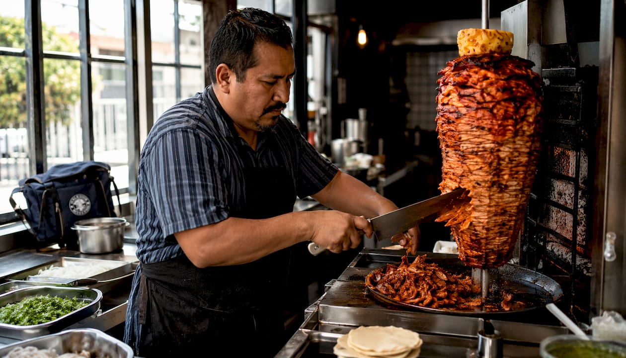 Un chef prepara y corta tacos al pastor en una cocina profesional, listo para servirlos bien calientes.