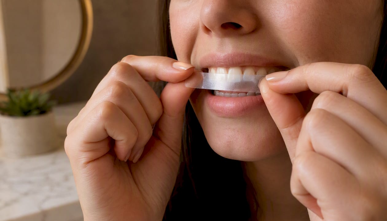 Une personne en train de poser une bande blanchissante sur ses dents.
