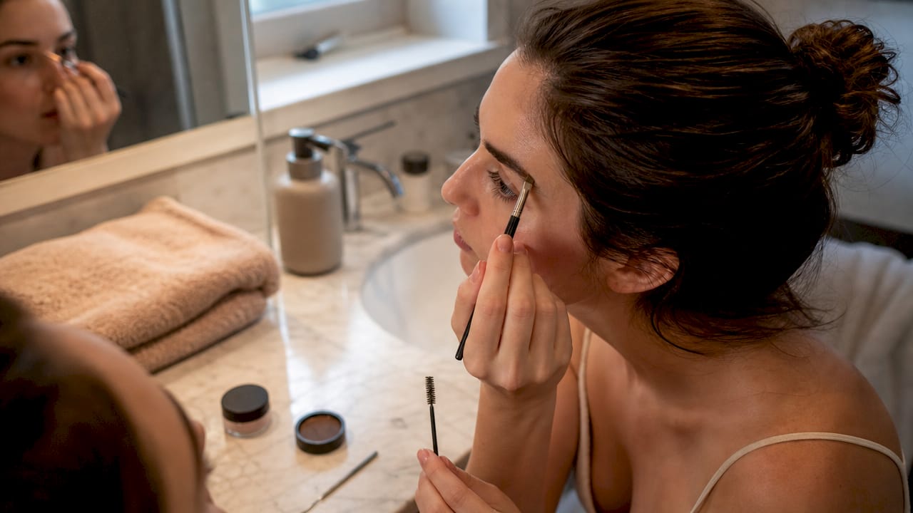 Manos maquillando las cejas con sombra, usando una brocha y un cepillo para darles forma y definición.