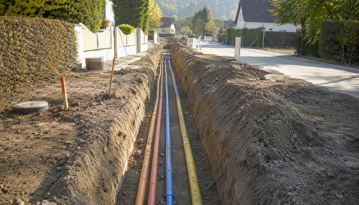 Ein Graben mit mehreren Versorgungsleitungen, in dem verschiedene Rohrsysteme für Strom, Wasser und Telekommunikation verlegt werden.