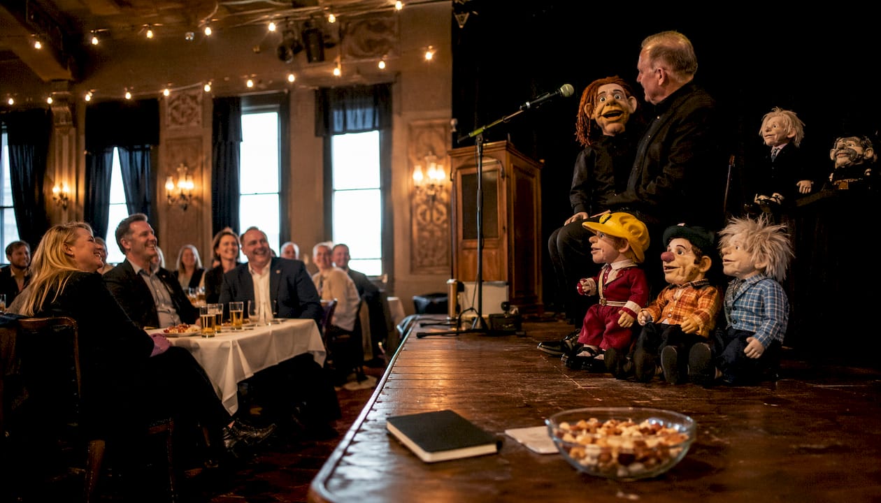 Underholdende firmaarrangement med bugtaler og sjove dukker på scenen, hvor publikum bliver en aktiv del af oplevelsen.