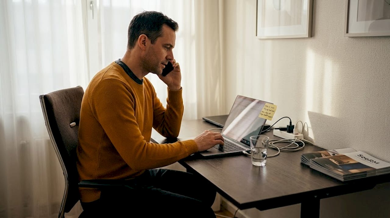 En man sitter vid skrivbordet och pratar i telefon samtidigt som han arbetar på sin laptop.