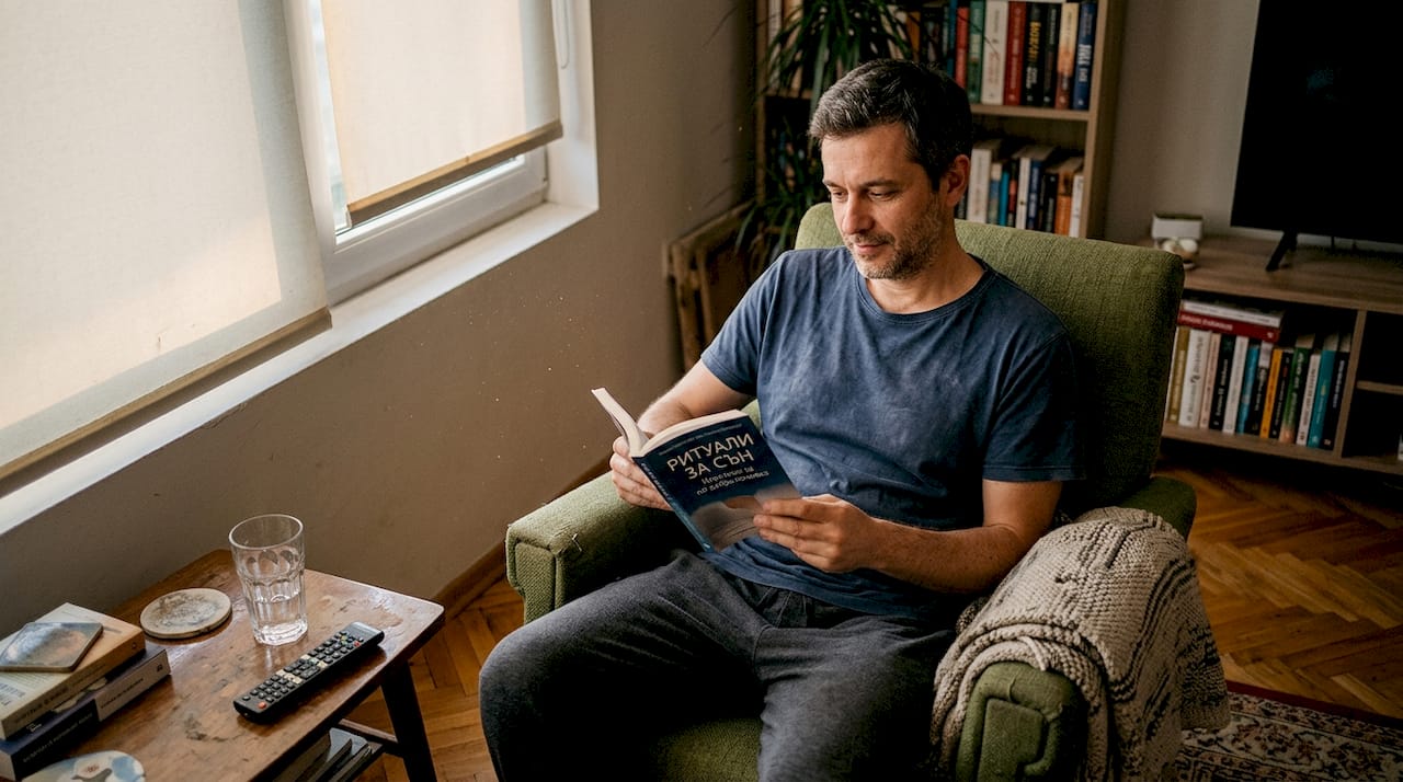 Мъж си почива вечерта в хола с книга в ръка, преди да си легне.