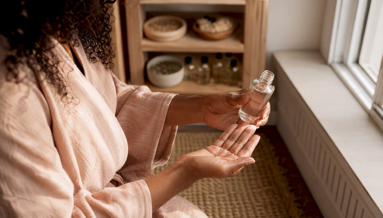 Et par hænder holder en flaske glycerin-hårserum i et lyst og indbydende badeværelse.