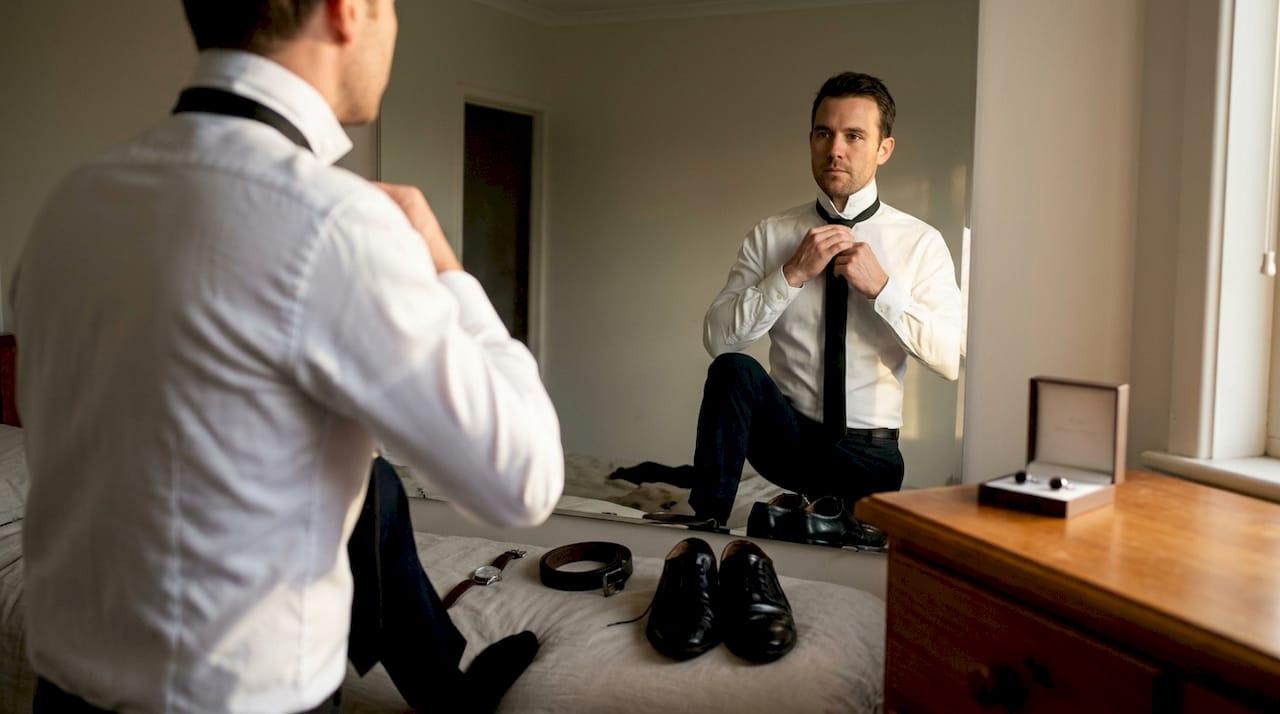 Man accessorizing budget formal outfit in bedroom