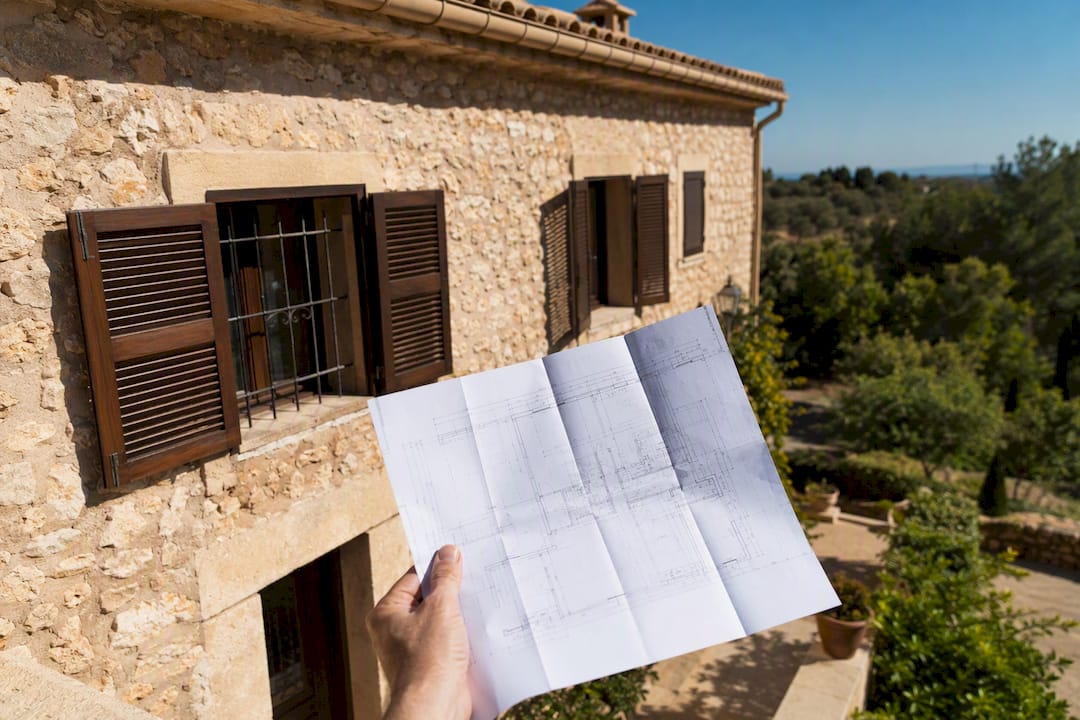 Hand hält Bauplan vor der Fassade einer Finca