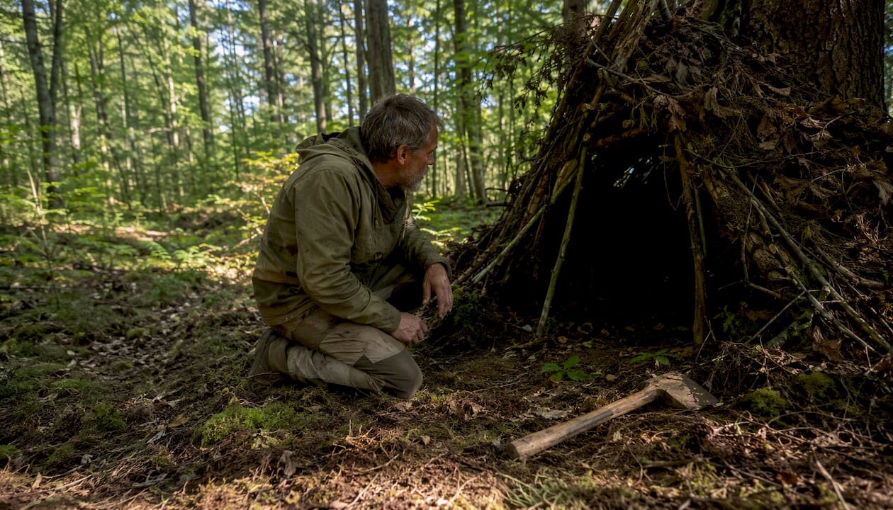 Man inspecting primitive debris hut outdoors