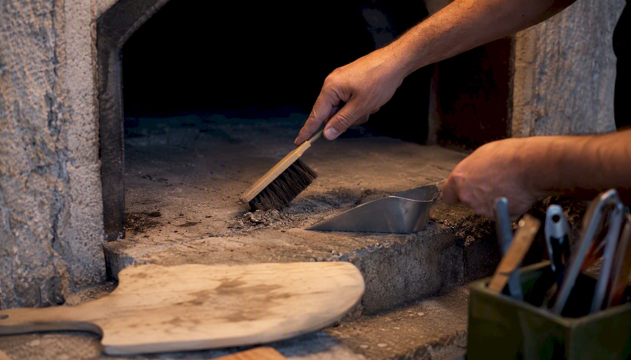 Mani che spazzano la cenere dal piano cottura caldo di un forno pizza