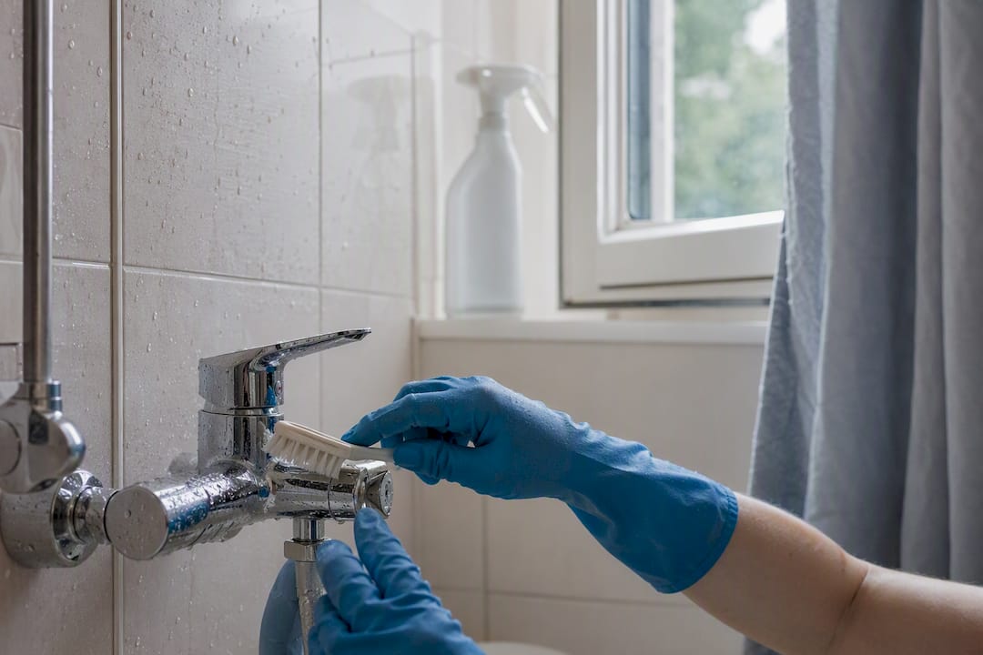 Ett par händer skrubbar rent fogarna mellan kakelplattorna i badrummet med en borste.
