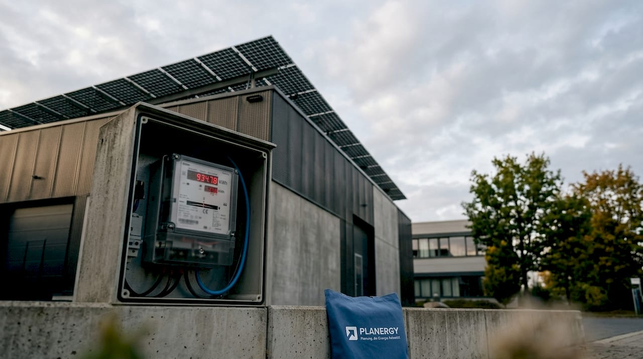 Im Mittelpunkt steht ein modernes Industriegebäude, dessen Dach mit Solarpaneelen ausgestattet ist. Der Energieverbrauch wird dabei präzise über einen digitalen Zähler überwacht.