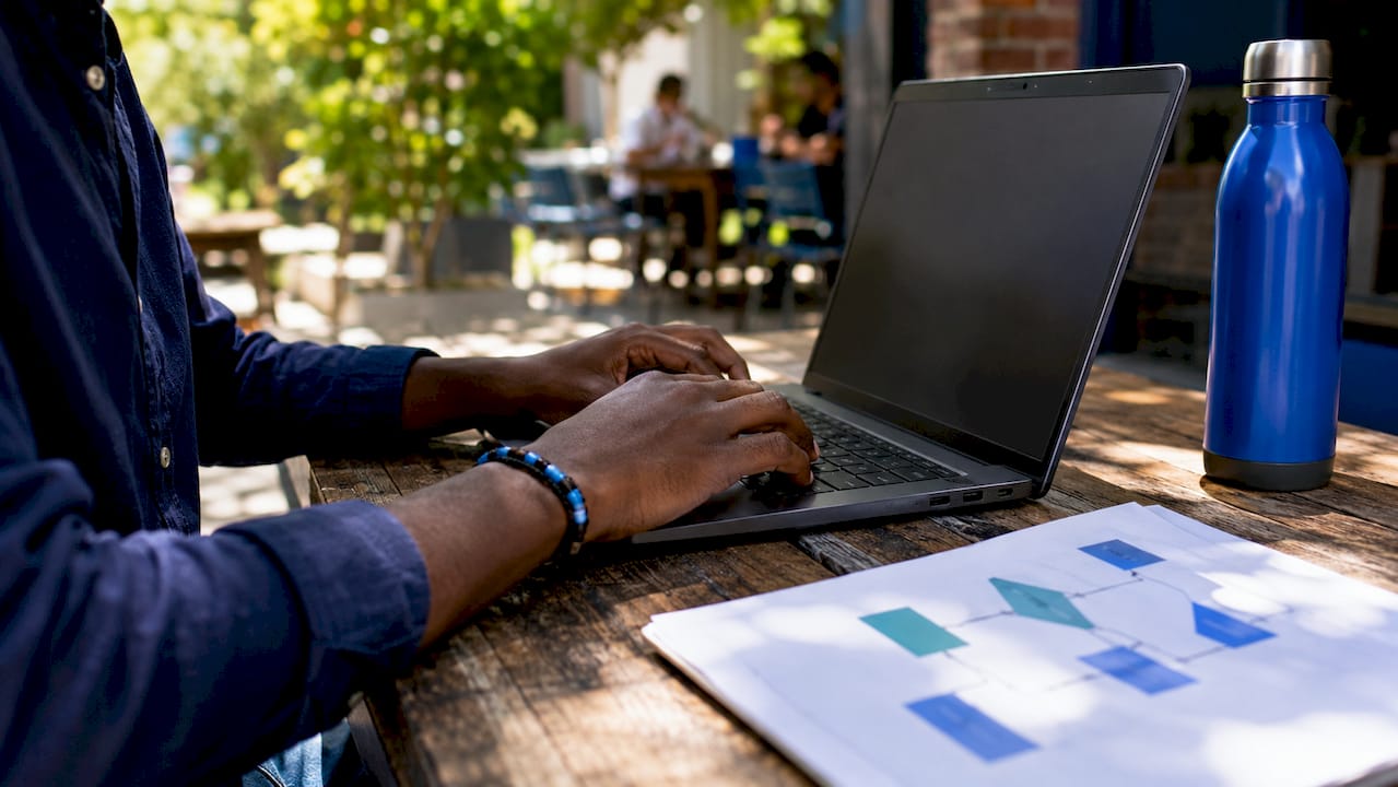 Hands typing on laptop with workflow papers on desk