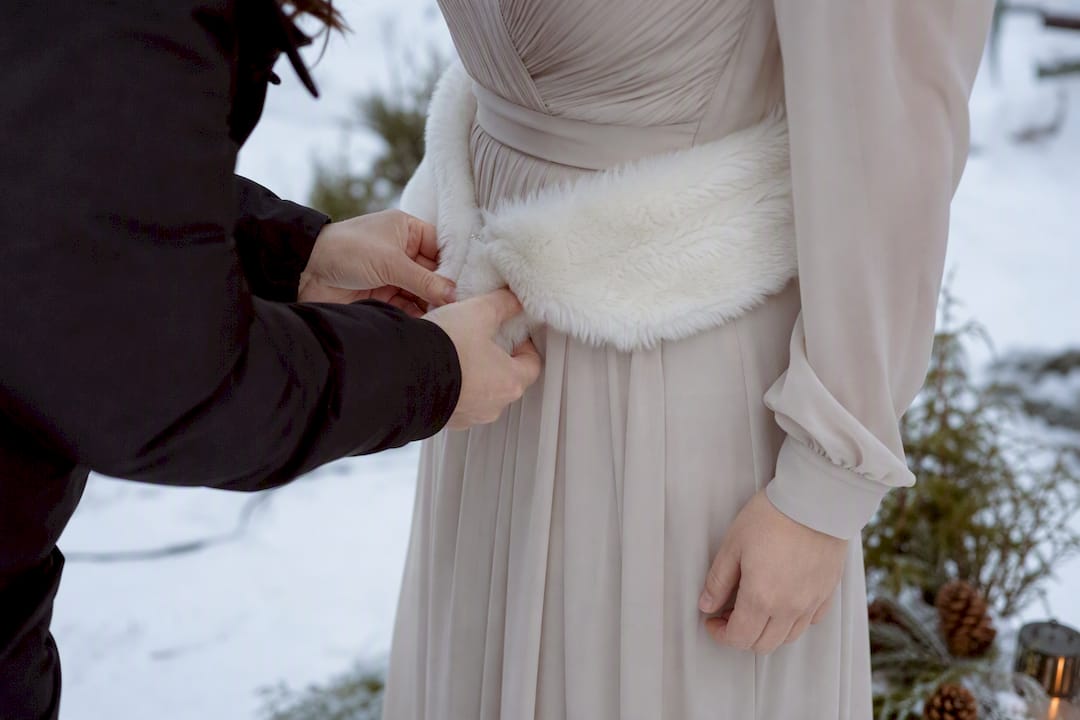 Bride’s hands tying fur sash at snowy ceremony
