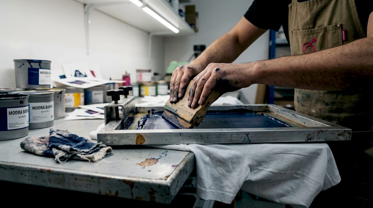 Na fotografiji so od blizu prikazane roke, ki nazorno prikazujejo postopek sitotiska.