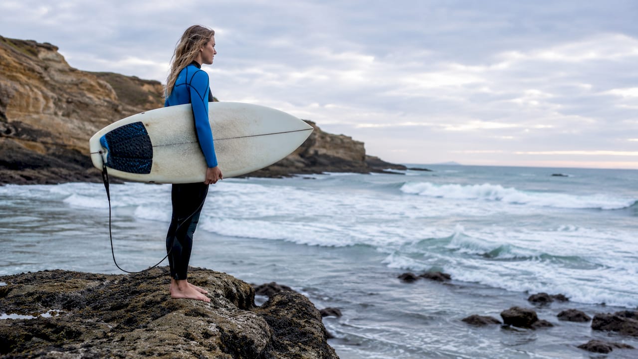 Woman surfer sitting on beach holding surfboard looking at waves