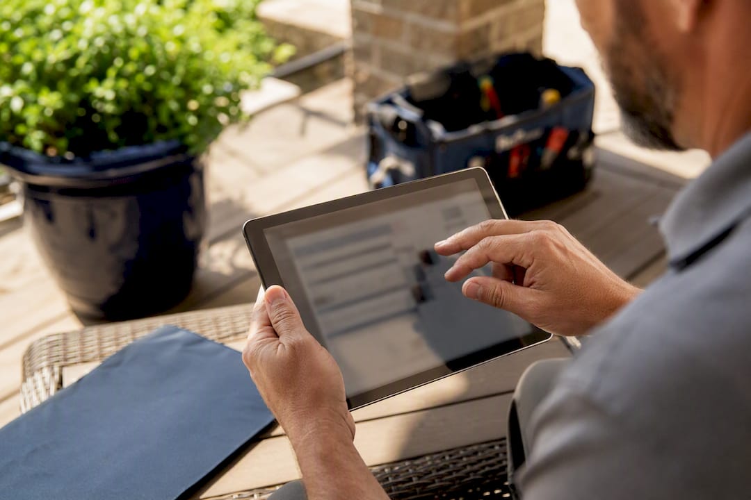 Man in a gray work shirt reviewing a claim estimate on a tablet at a patio table, with a tool bag nearby