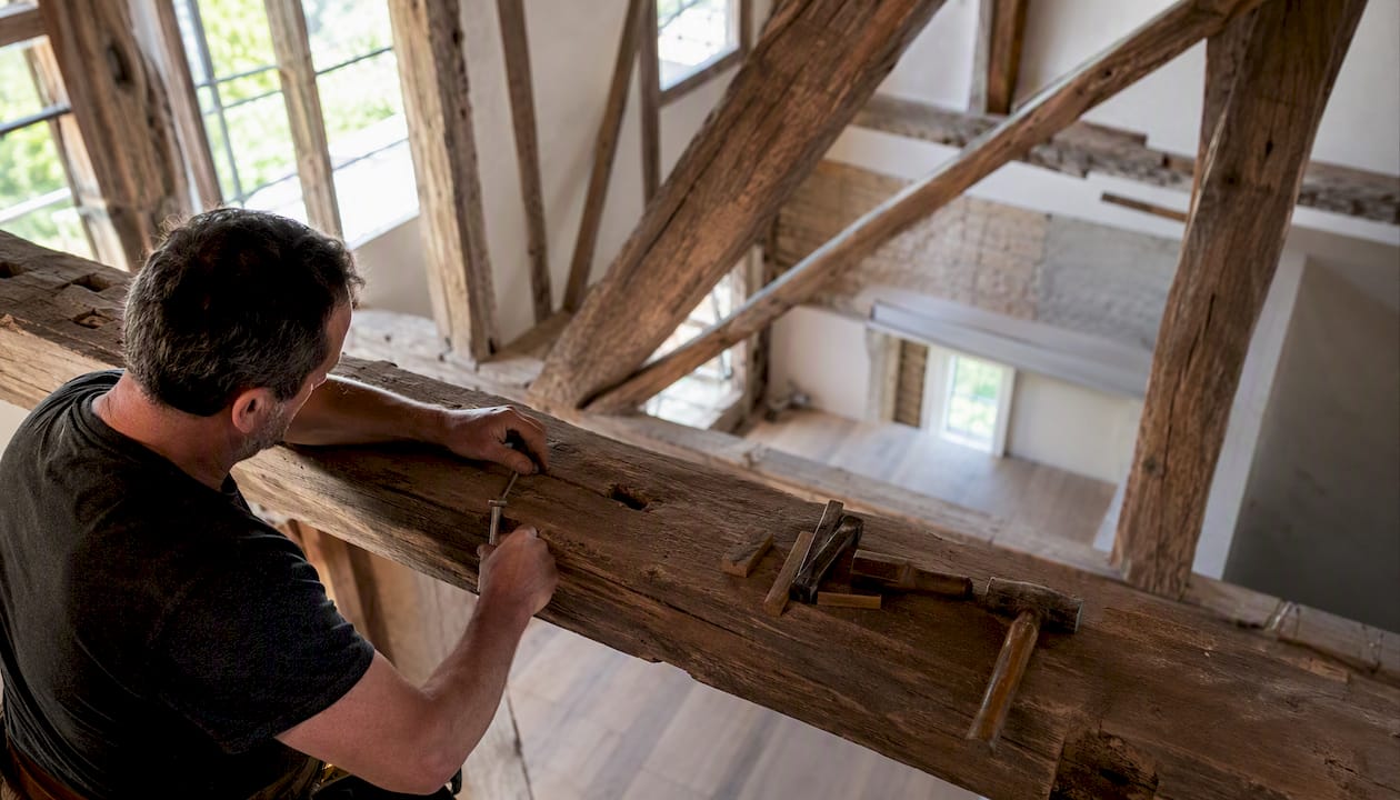 Un charpentier ajuste une poutre sur la toiture, guidant le bois avec précision grâce à son savoir-faire.