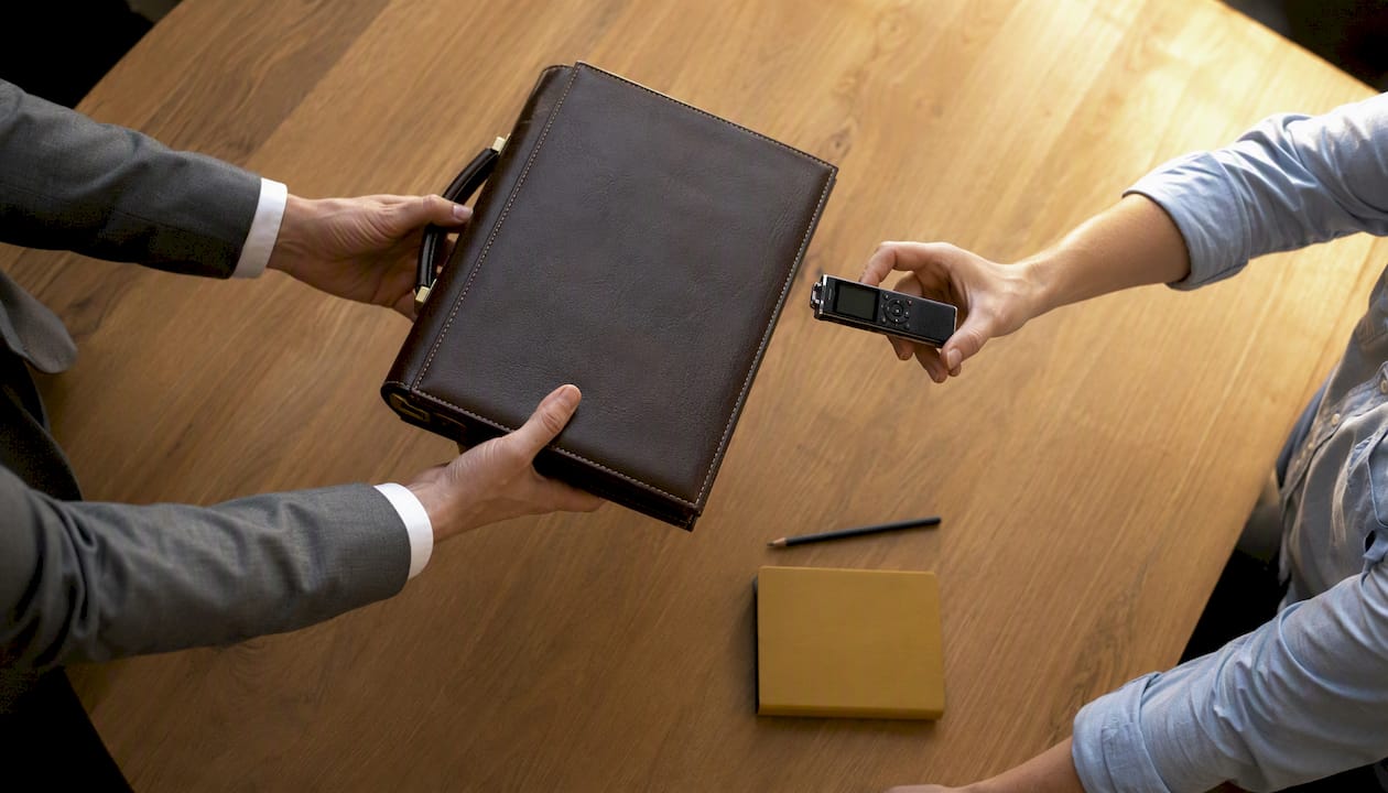 Au cours d’une réunion professionnelle, des mains s’échangent un porte-documents et un enregistreur vocal.