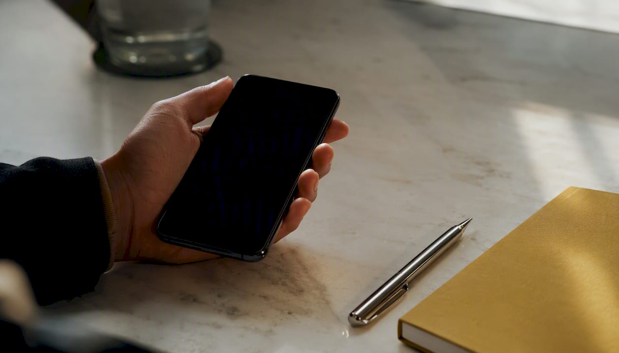 Une main tenant un smartphone au-dessus d’un bureau parsemé d’accessoires de travail.