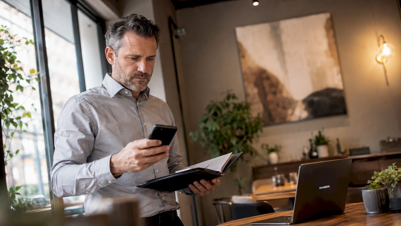 Ein Mann sichtet im Büro Unterlagen und checkt sein Smartphone.