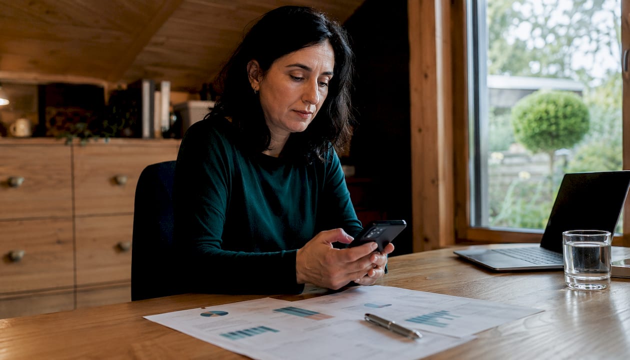 Eine Frau arbeitet mit ihrem Smartphone und hat technische Unterlagen vor sich liegen.