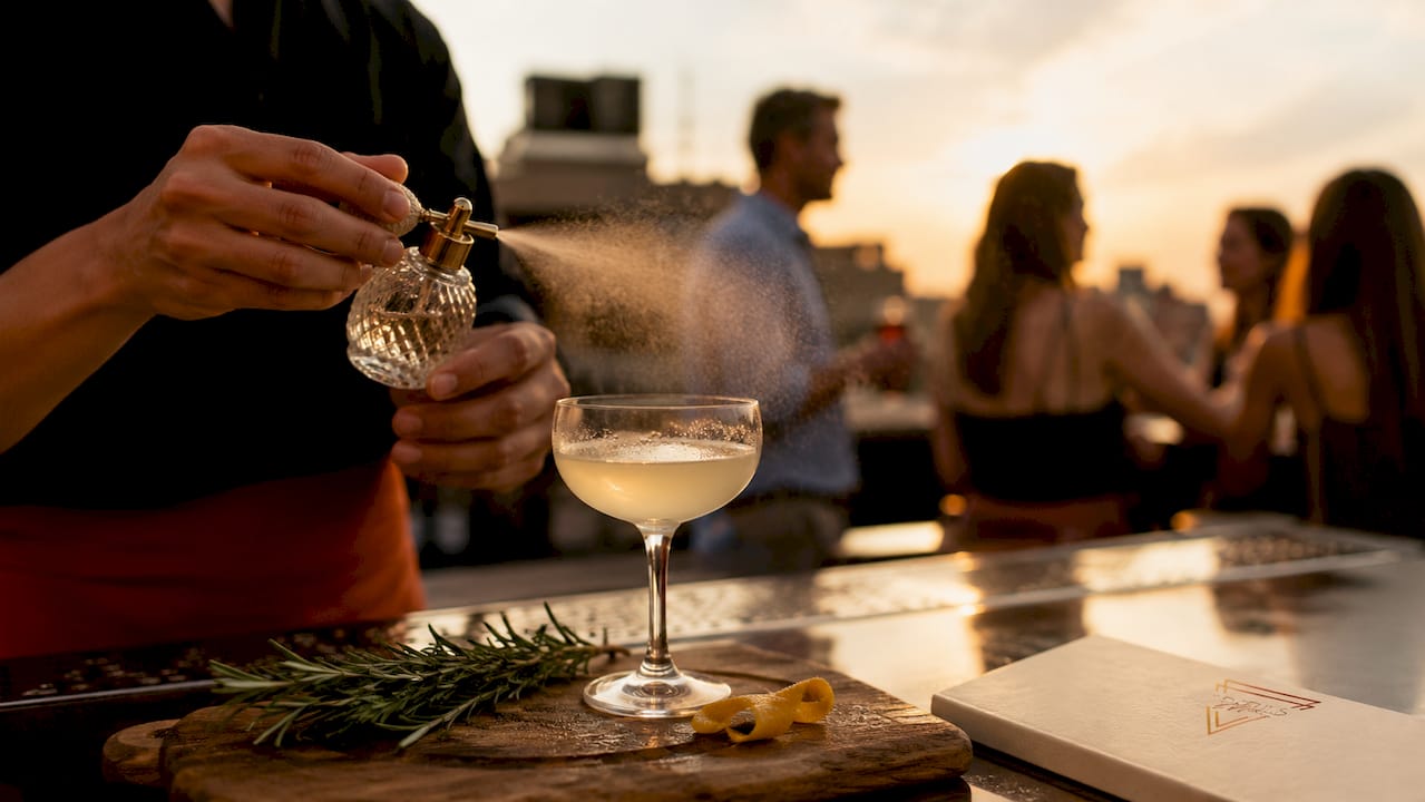 Twee handen vernevelen een kruidige geurwolk boven een cocktailglas.