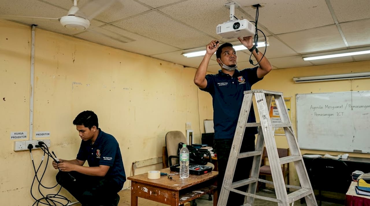 Seorang juruteknik sedang memasang projektor di bahagian siling dalam sebuah bilik.