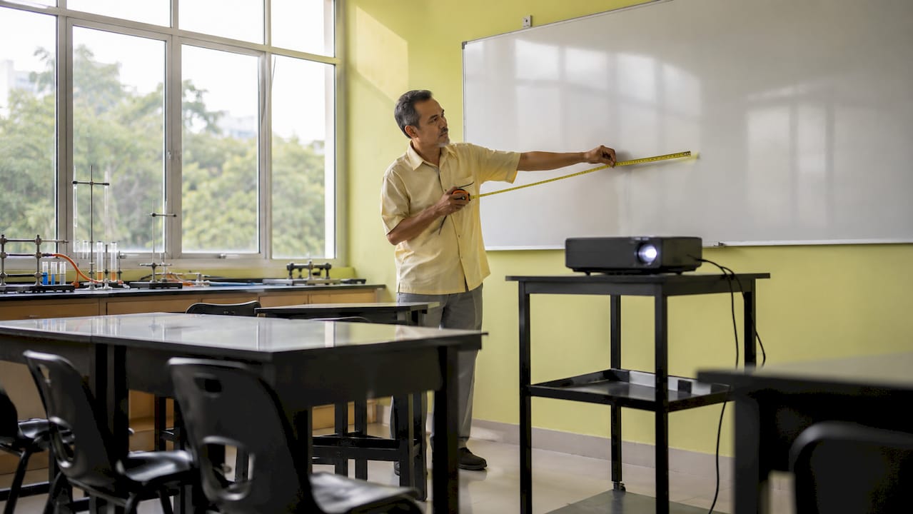 Guru sedang mengukur jarak projektor di dalam kelas bagi memastikan paparan jelas untuk semua pelajar.