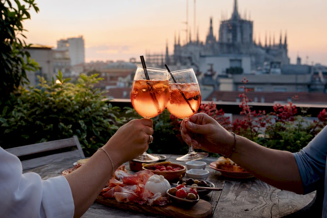 Hands holding Spritz aperitivo glasses on Milan rooftop at sunset