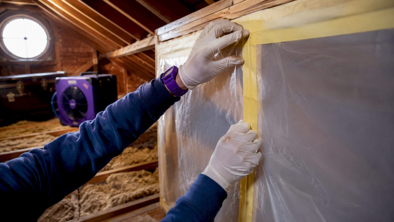 Hands sealing plastic containment barrier in attic