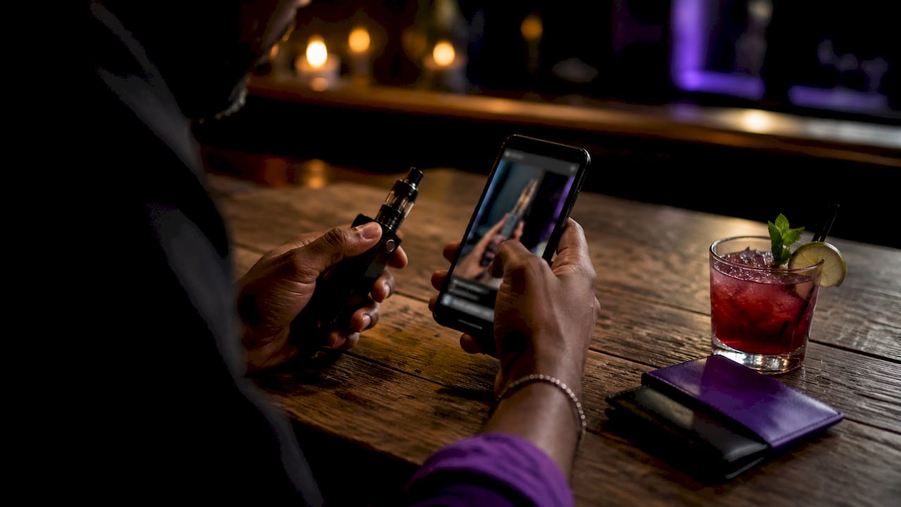 Hands holding vape and smartphone with reviews in café