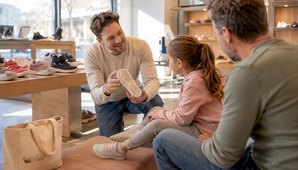 Ein Verkaufsberater zeigt einem Kunden am Tresen eines Sportgeschäfts die Besonderheiten der Sohle eines Kinderschuhs und erklärt, worauf beim Kauf zu achten ist.