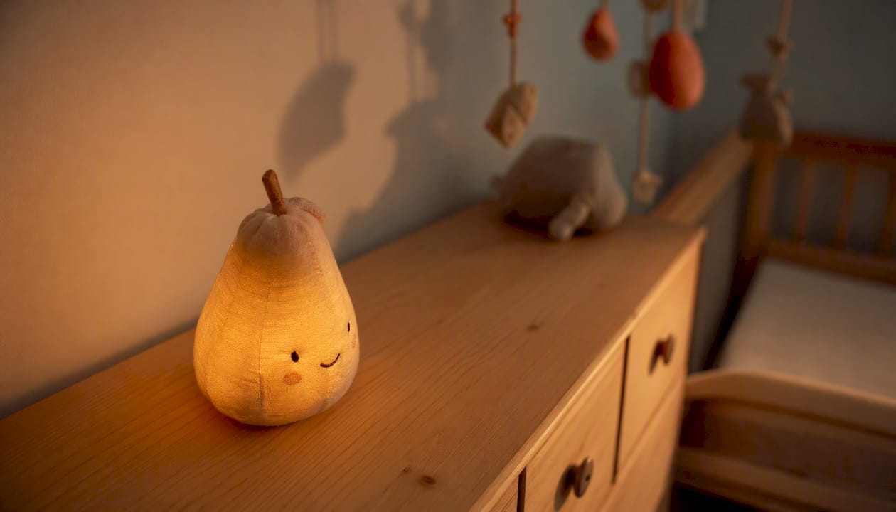 Une veilleuse diffuse une lumière douce sur la commode en bois, enveloppant la pièce d’une ambiance apaisante pendant la nuit.