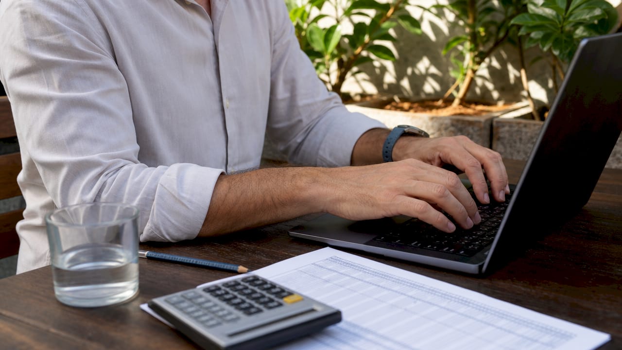 Persona al lavoro al computer, con calcolatrice e fogli sparsi sulla scrivania.