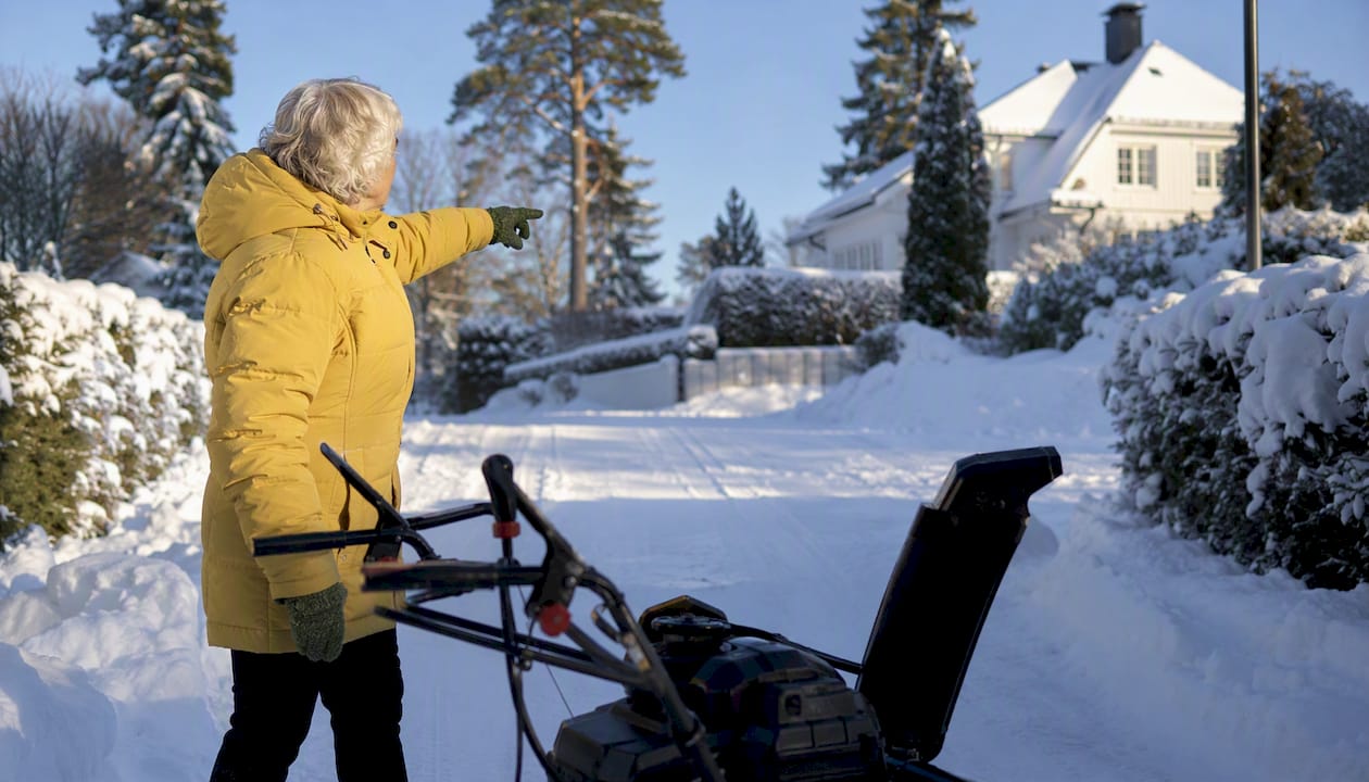 Äldre man skottar snö med snöslunga och ser nöjd ut i trädgården.