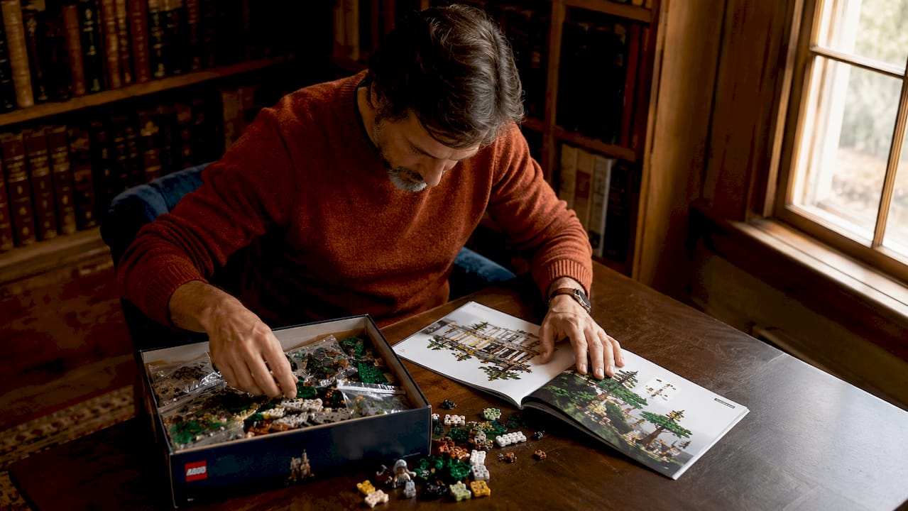 Un homme installé à une table examine un set LEGO tout en feuilletant le catalogue, cherchant à comparer les modèles.
