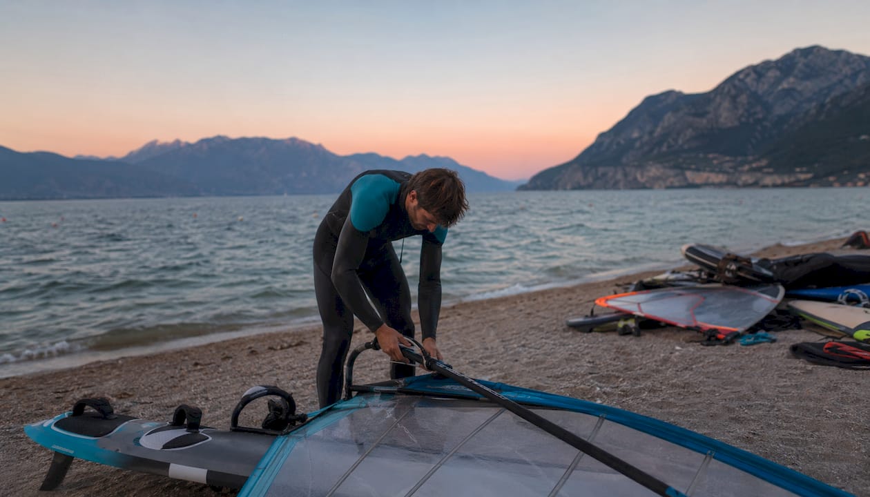Un windsurfista si sta preparando per entrare in acqua sul Lago di Garda, sistemando la sua tavola e la vela sulla riva.
