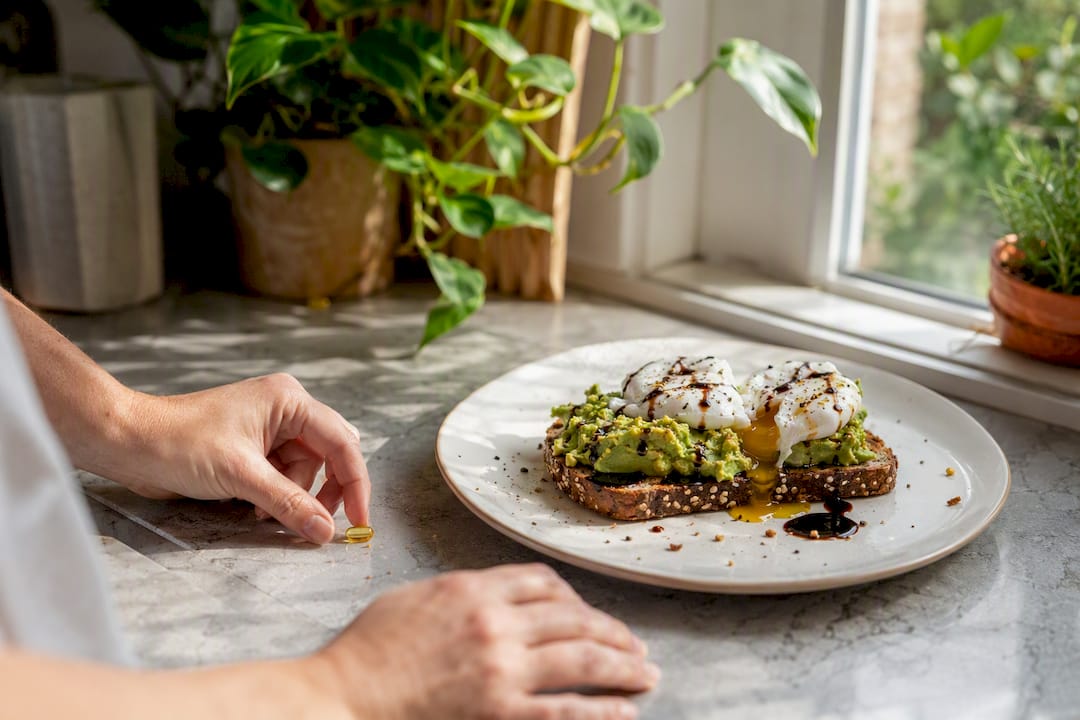 Hand placing D3 K2 softgel beside avocado toast and egg