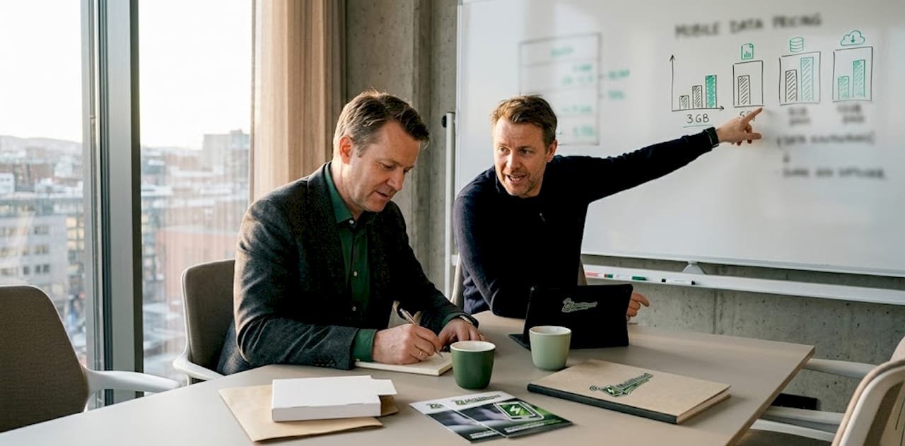 To menn sitter rundt et møtebord og diskuterer ulike mobilabonnement.