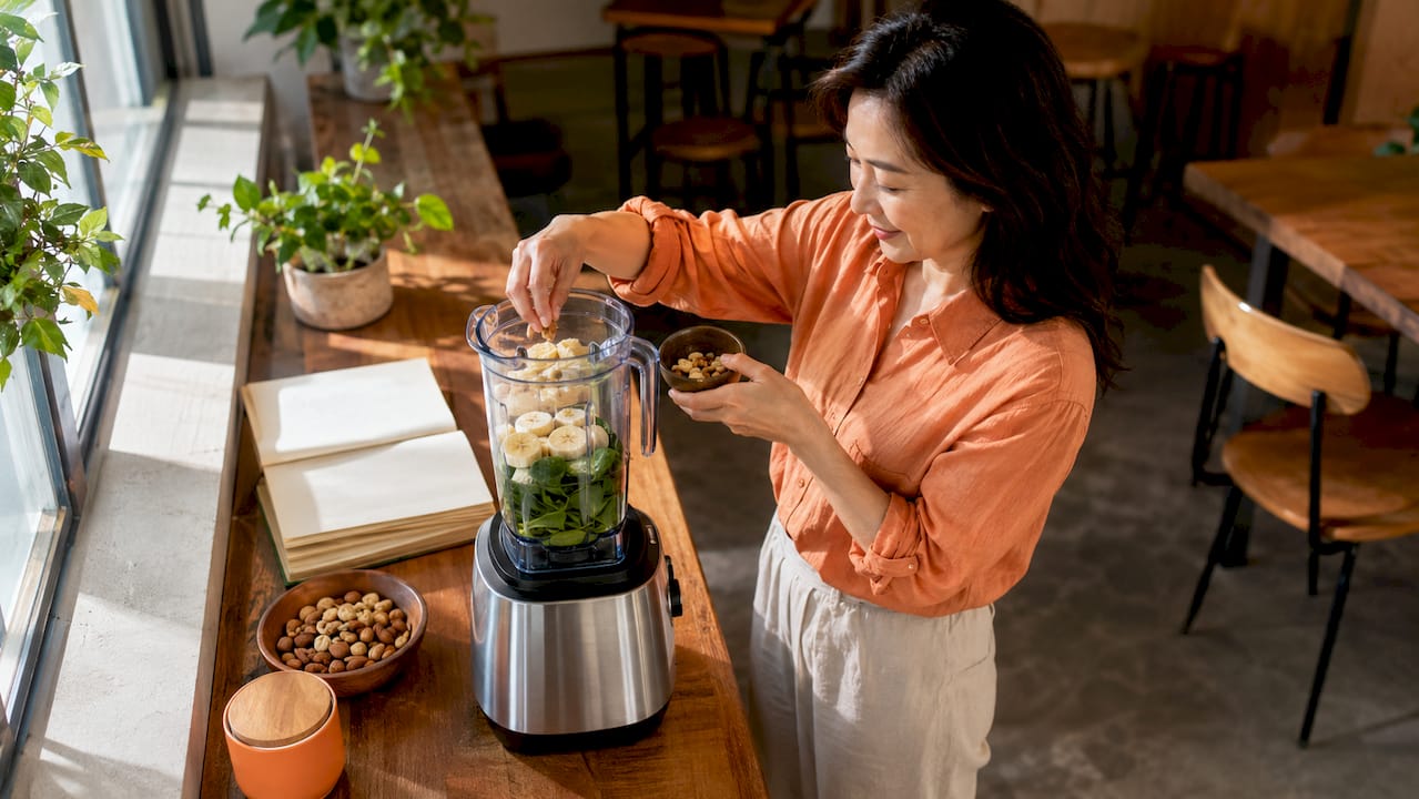 Woman preparing spinach banana smoothie in kitchen