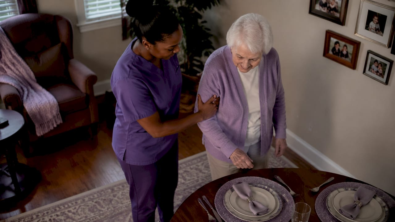 Home health aide helping elderly woman walk in kitchen