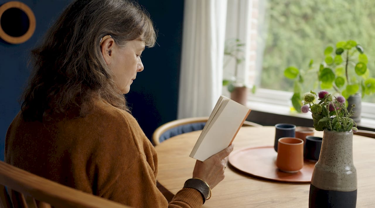 Een vrouw met een hoortoestel zit aan tafel en kijkt op haar smartphone.