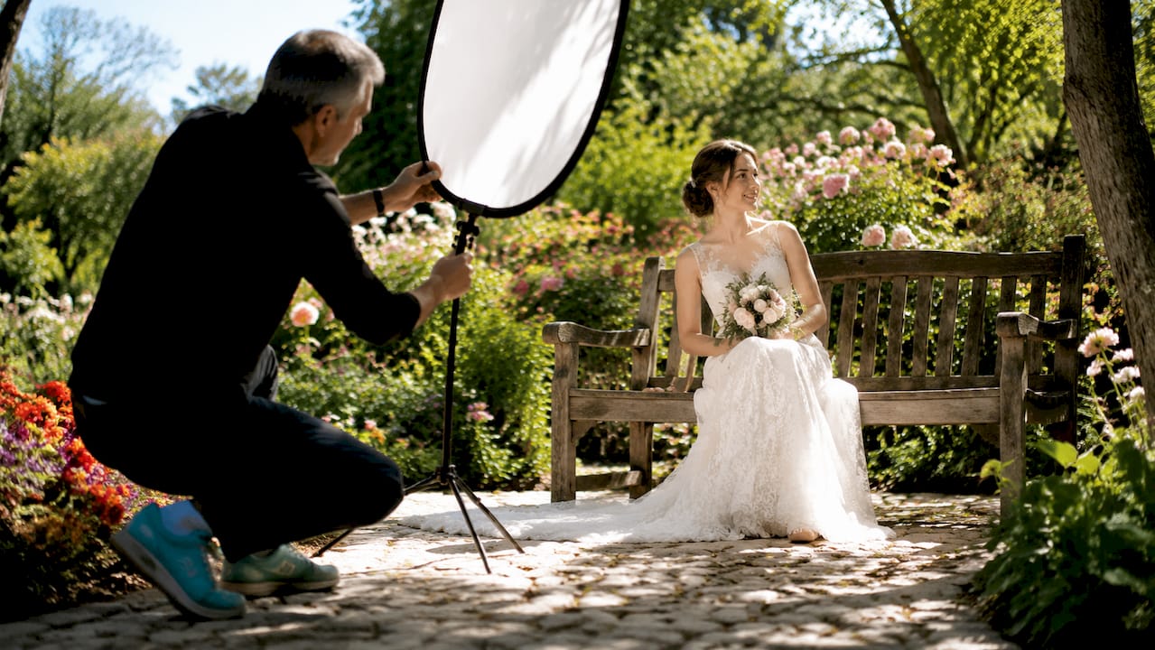 Photographer shoots a seated bride in a white dress on a garden bench, with bright flowers and greenery.