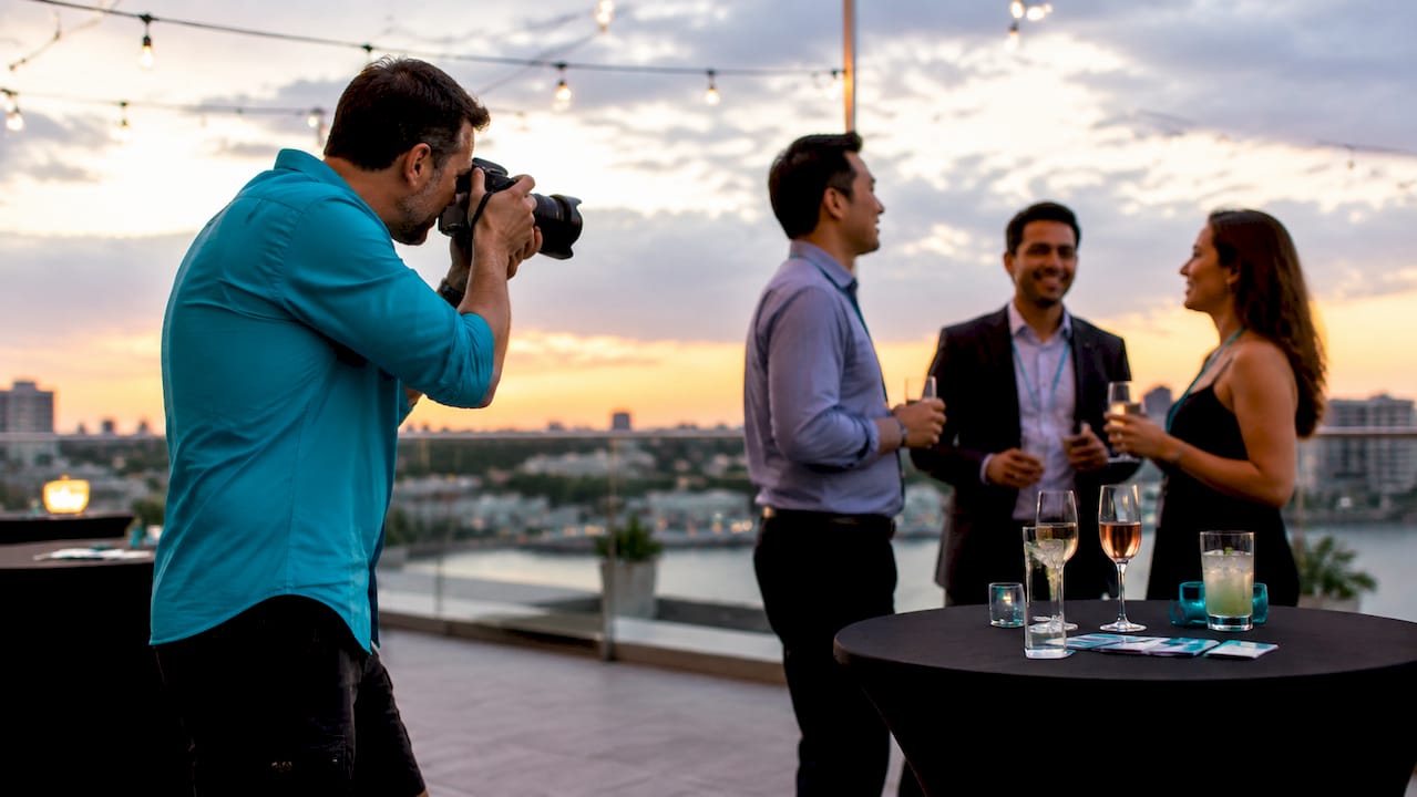 Photographer taking a rooftop sunset shot of three people chatting beside a table with drinks.