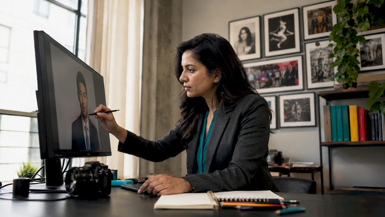 Person in a blazer works at a desk, pointing at a large monitor in a bright home office.