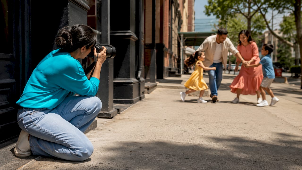 Photographer crouching on sidewalk, taking a photo of children running on a sunny street.
