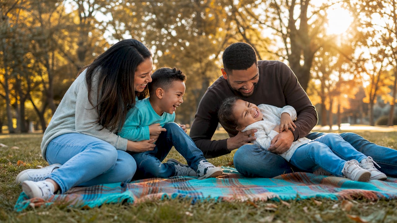 Family sitting on a blanket in a park, smiling and cuddling a baby at sunset